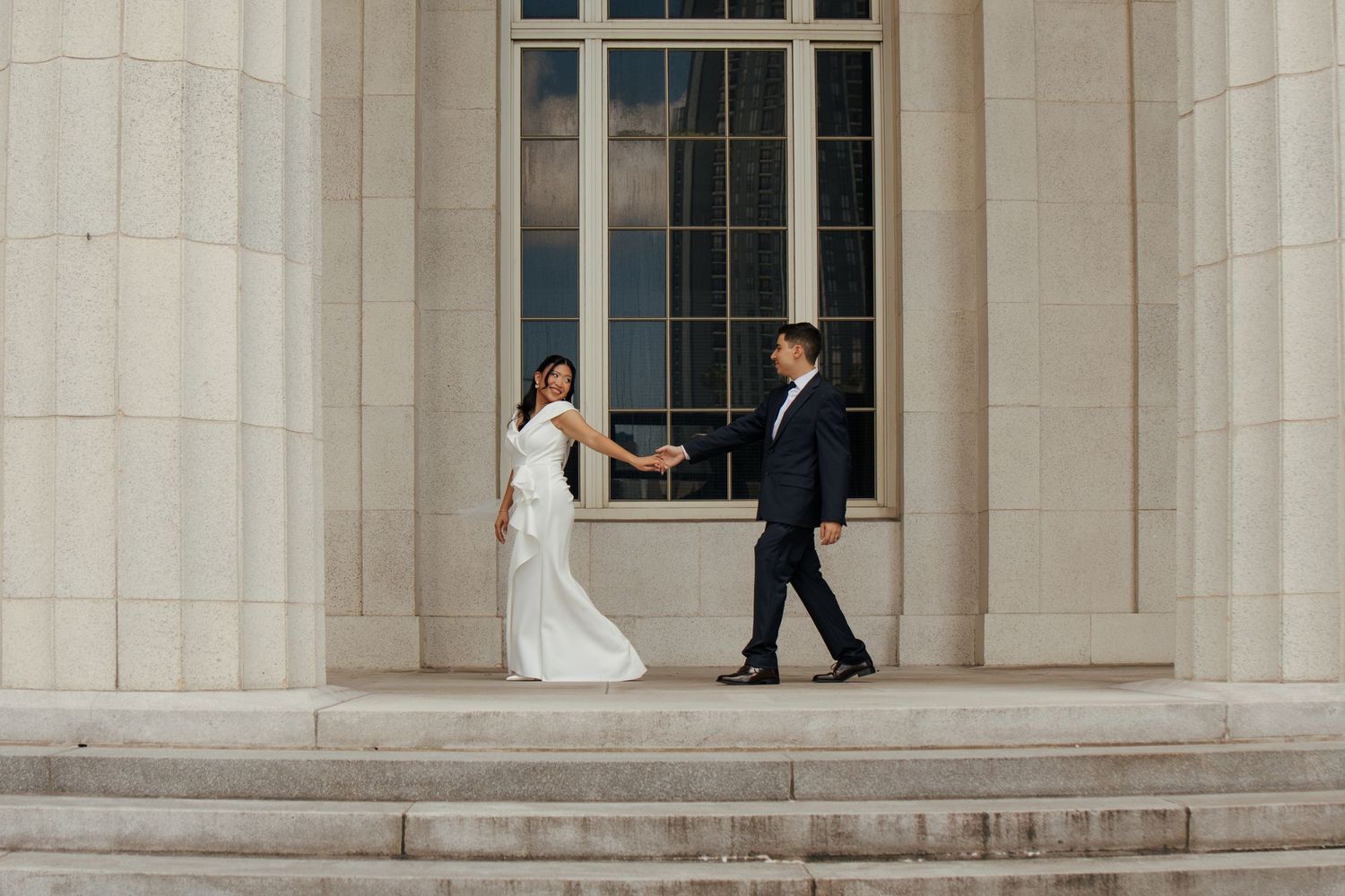 Bride and groom walk along stone steps between tall columns during outdoor wedding portraits.