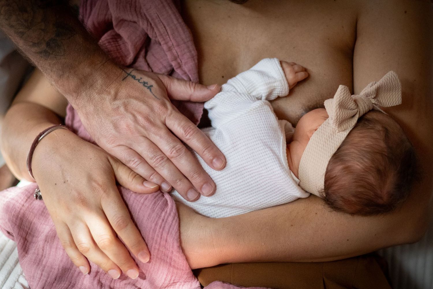 Parents cradling their swaddled newborn baby girl with a bow headband together.