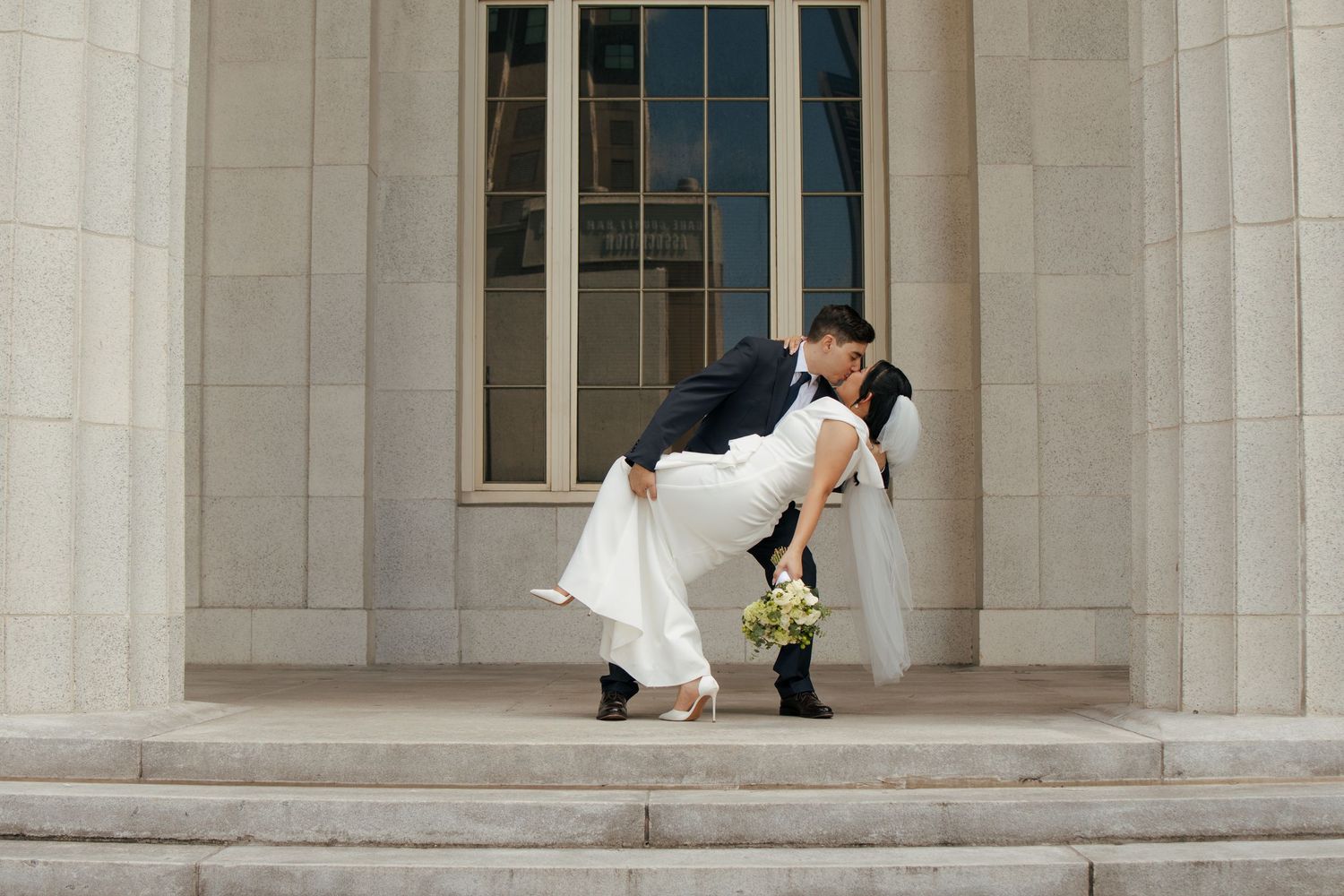 Groom dips bride in elegant white gown on stone steps during wedding portrait session.