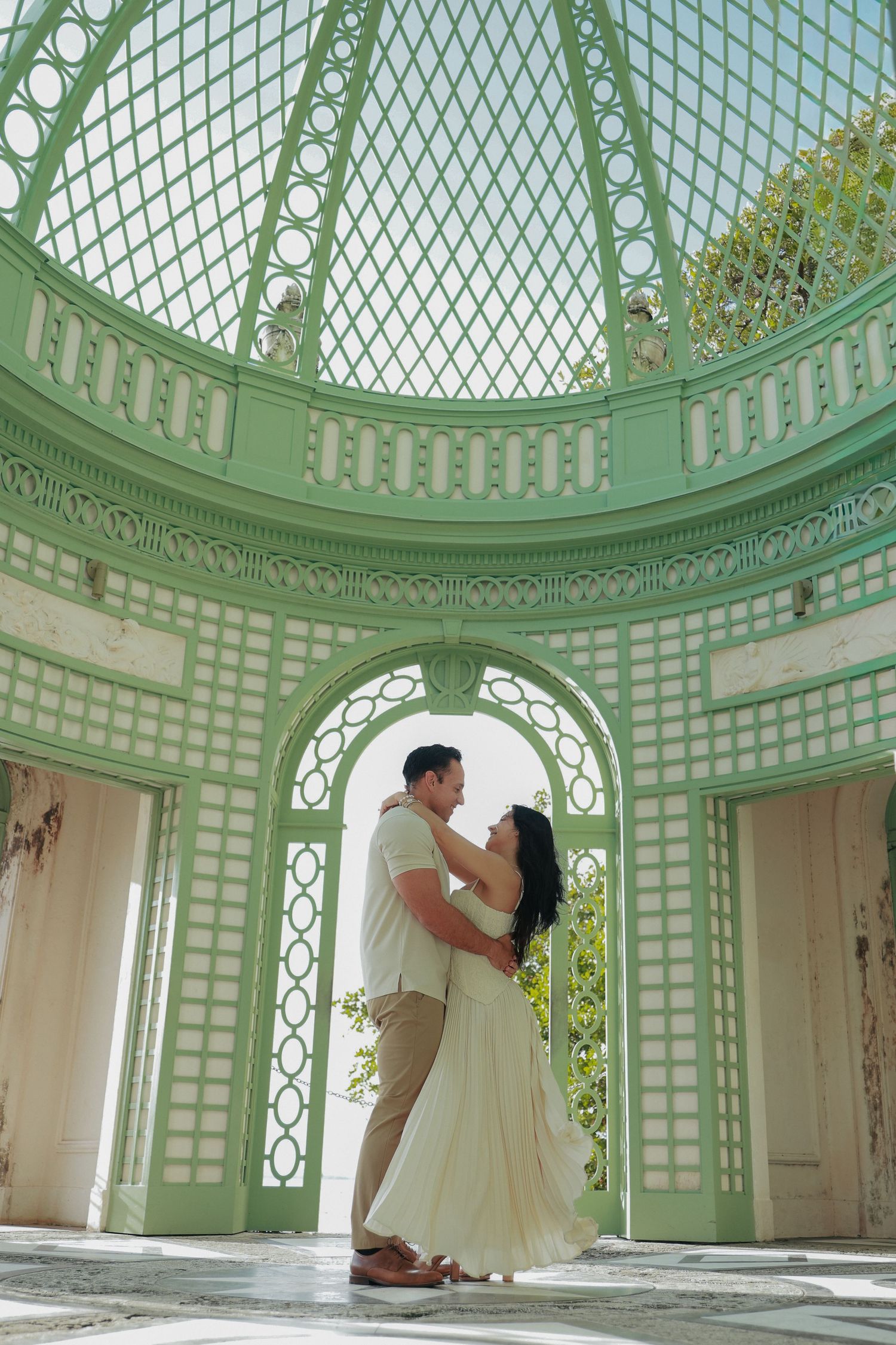 Couple embracing under ornate green lattice dome in elegant architectural setting.