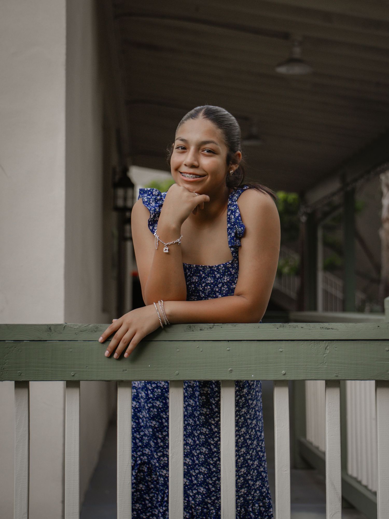 Smiling teenage girl in blue floral dress leaning on green railing of a porch.