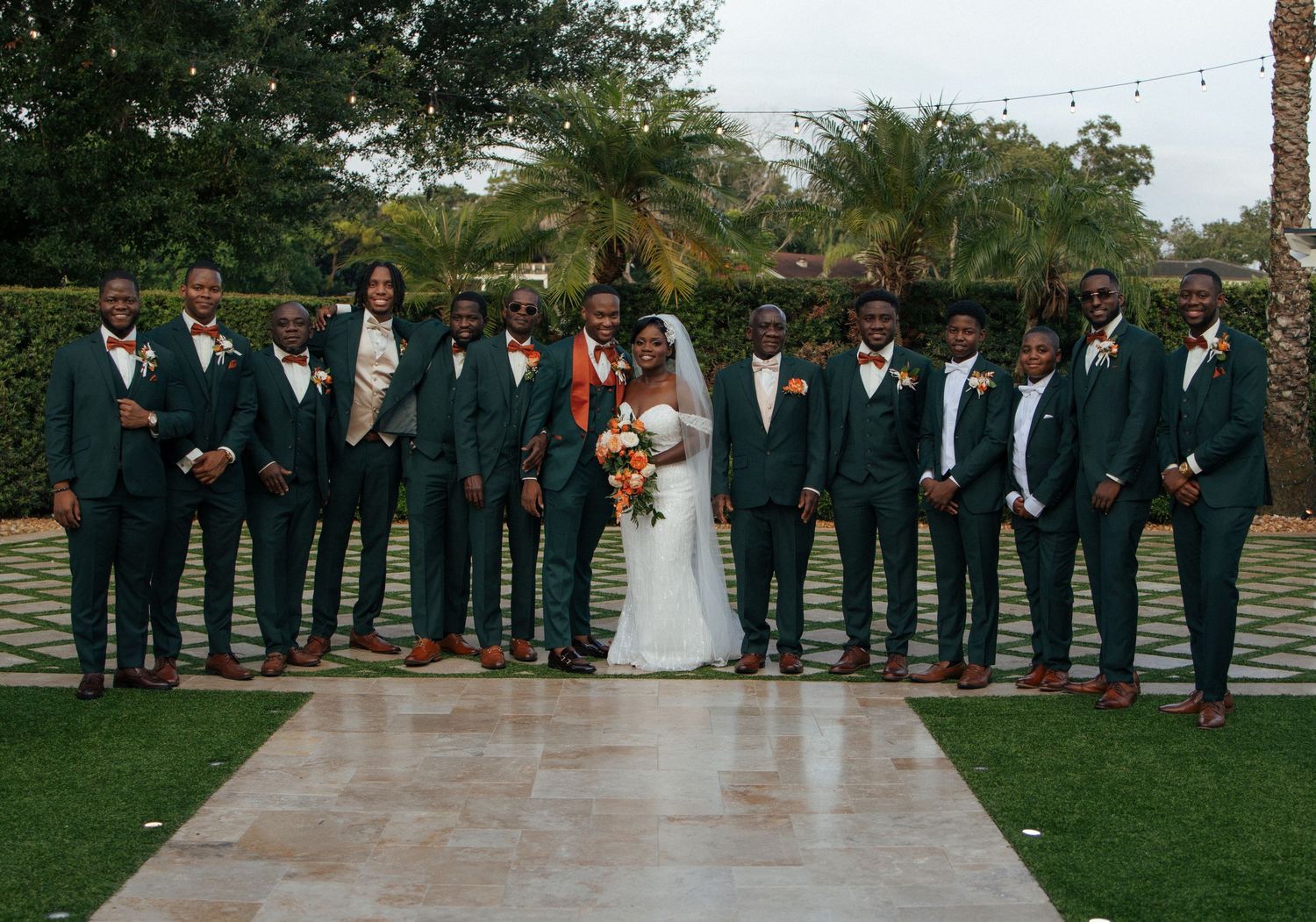 Full wedding party portrait outdoors with groomsmen in dark green suits and bride holding an orange bouquet.