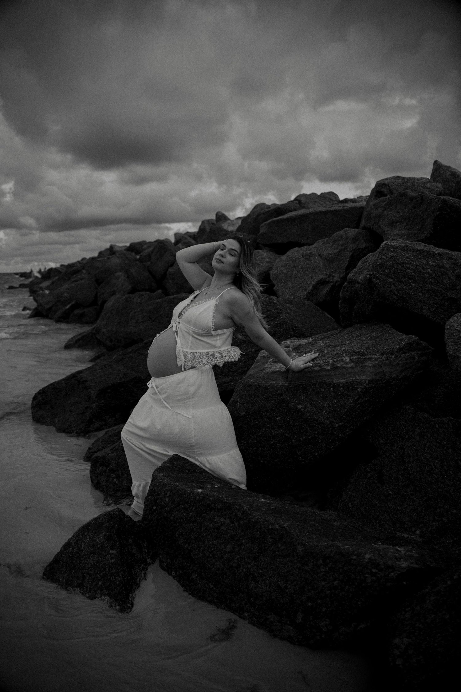 Black and white photo of pregnant woman in white dress posing on rocky shoreline with stormy sky.