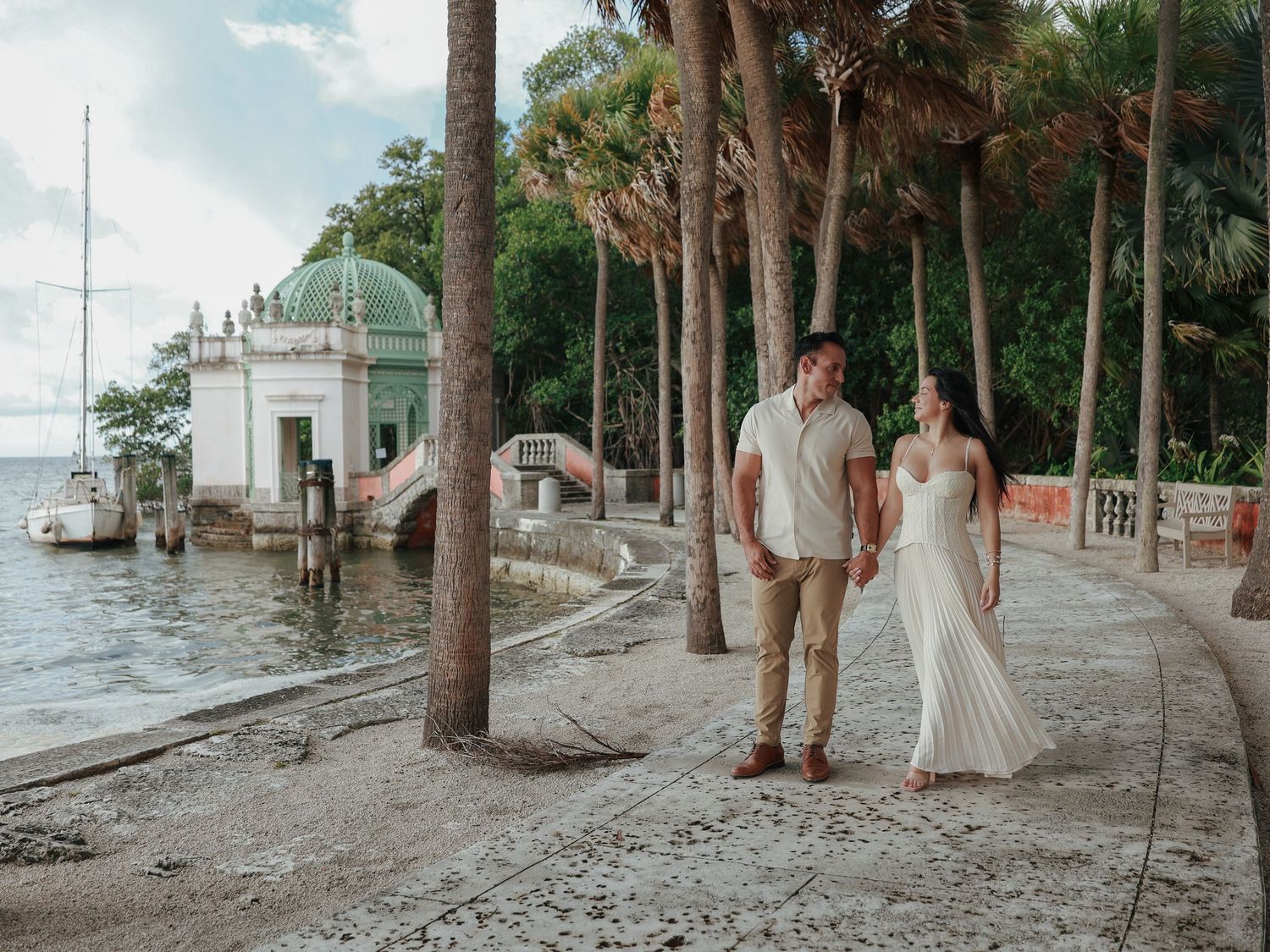 A couple walks hand-in-hand along a palm-lined waterfront path near a historic domed building.