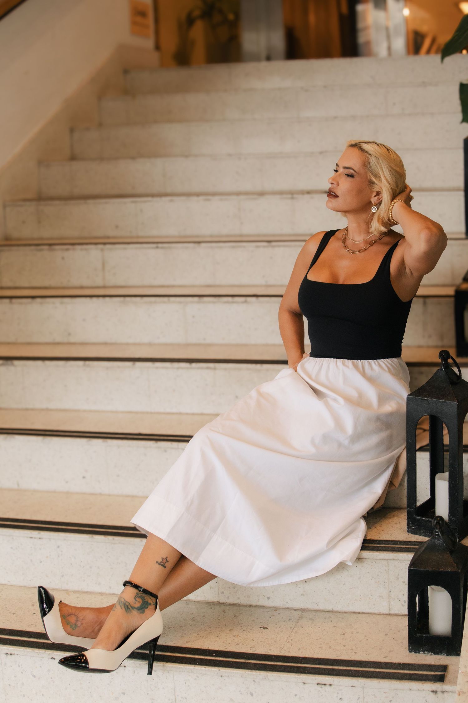 Stylish blonde woman sitting on marble stairs in white skirt and black heels with ankle tattoo.