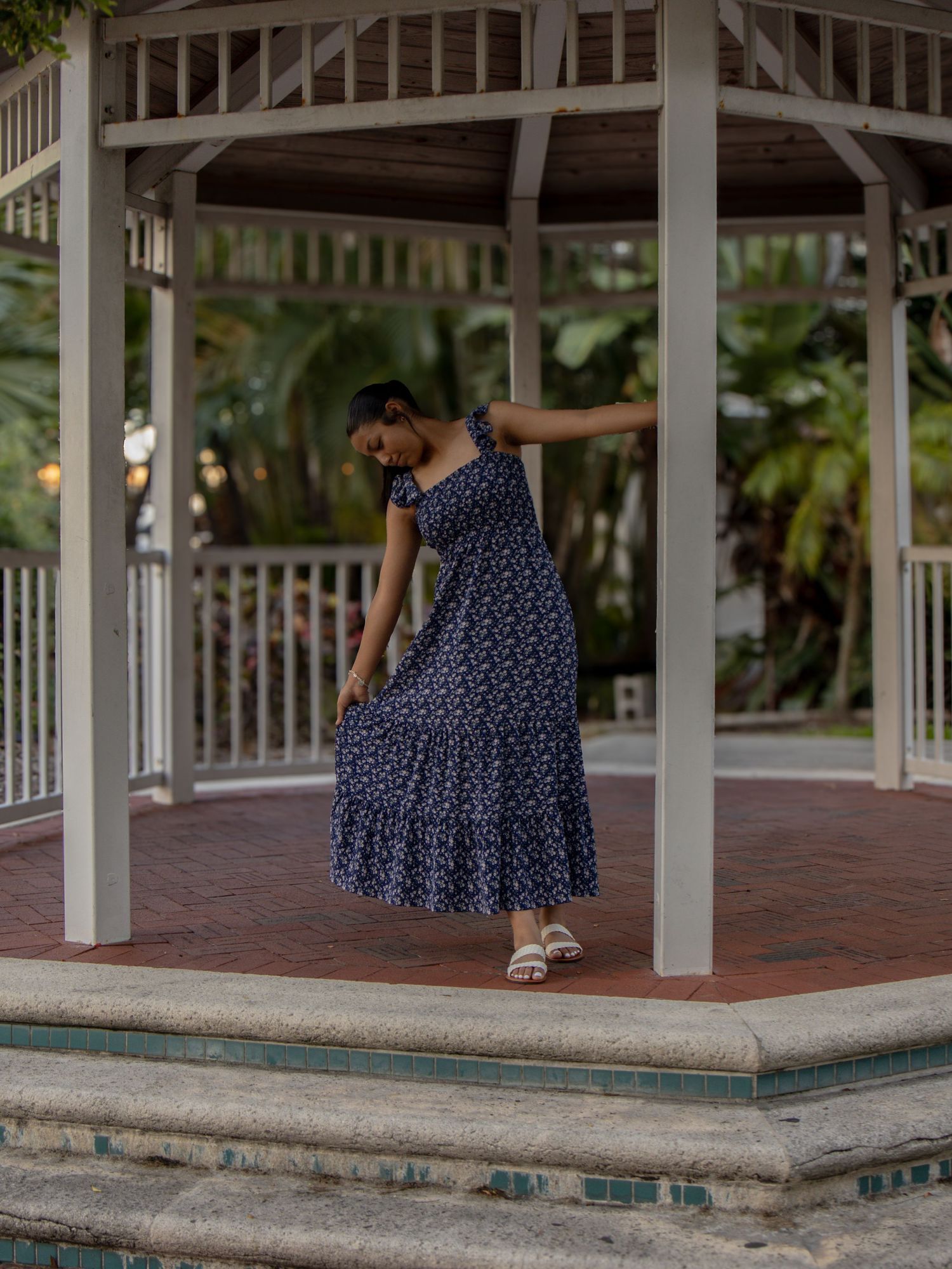 Young woman in navy floral maxi dress posing gracefully inside a white wooden gazebo.