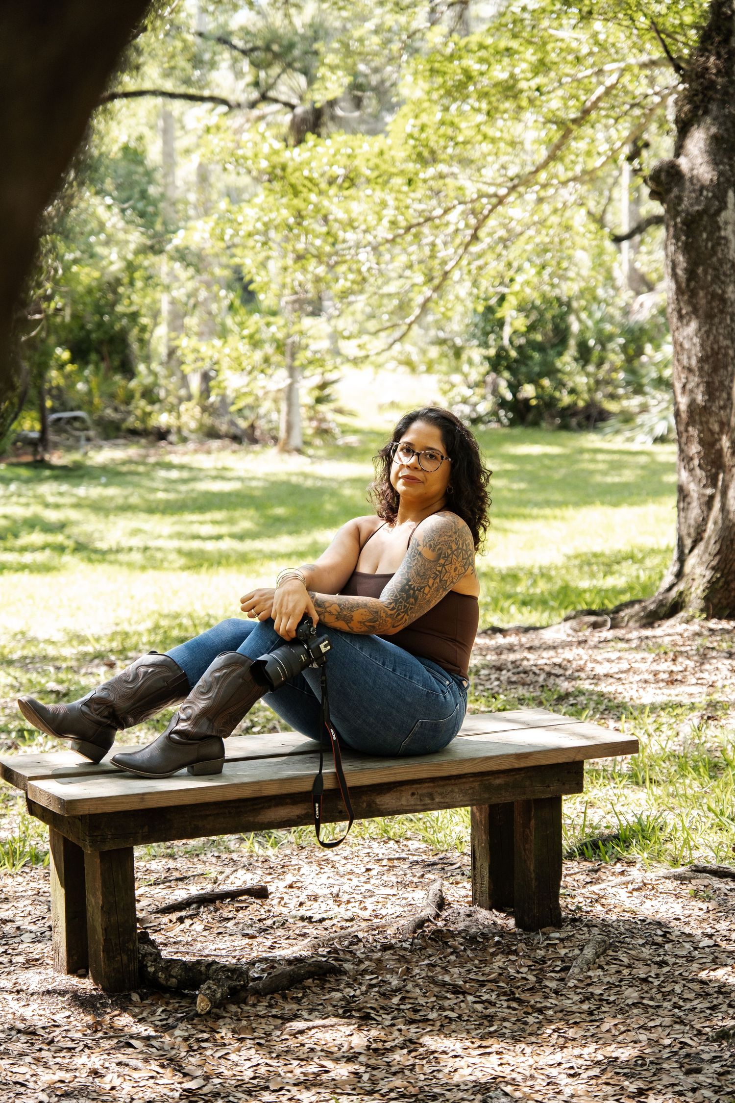 A tattooed woman in jeans and boots sits relaxed on a wooden bench in a sunlit park.
