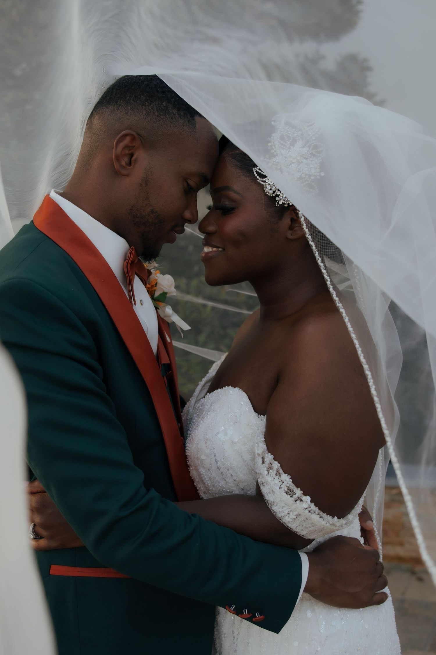 A bride and groom share a tender forehead-to-forehead moment under a flowing veil outdoors.
