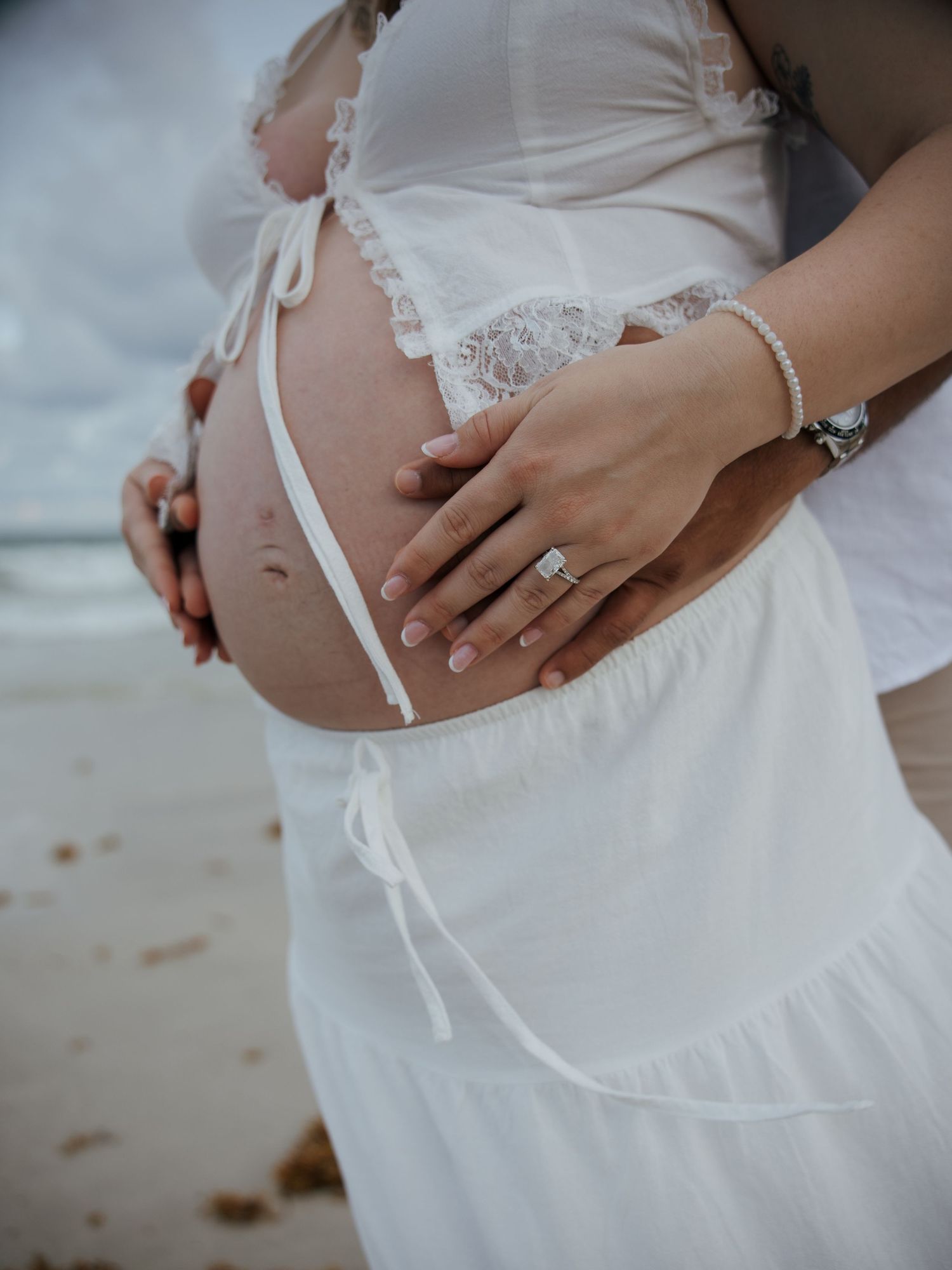 Close-up of pregnant belly with couple's hands cradling bump, woman wearing engagement ring and bracelet.