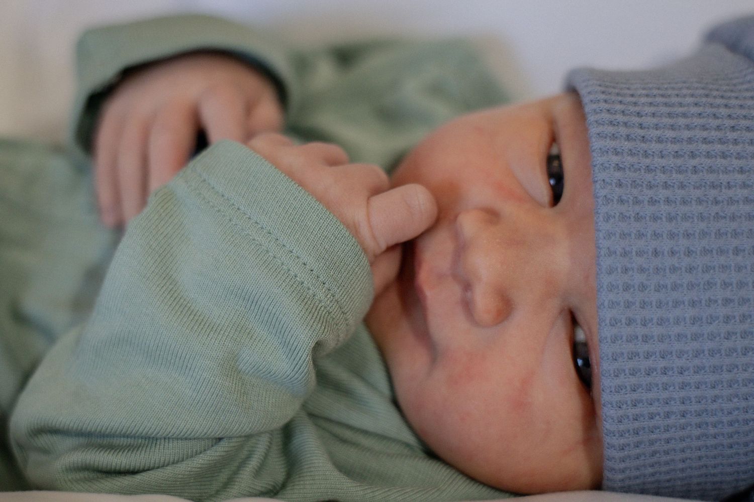 Newborn baby in green outfit and gray hat gently touching face with small hand.