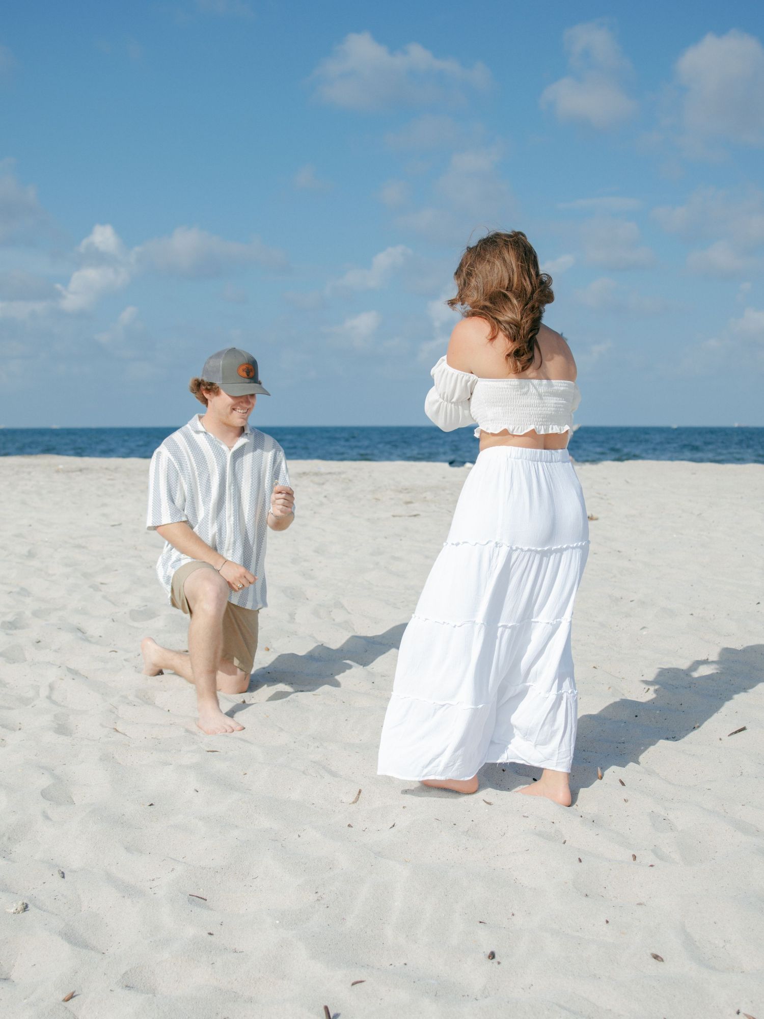 Couple on white sand beach, man kneeling before woman in white off-shoulder crop top and maxi skirt during proposal.