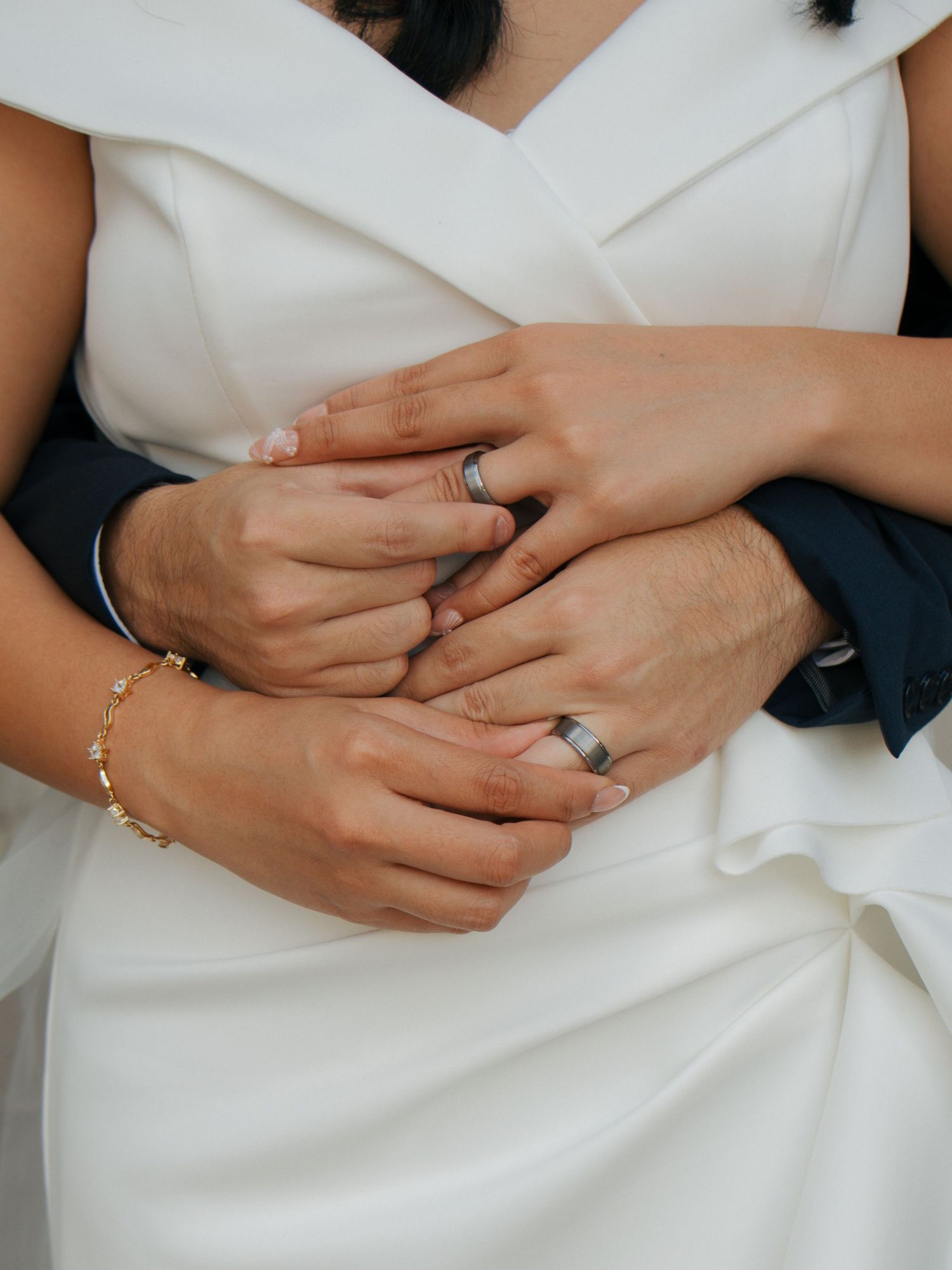 Close-up of bride and groom's interlaced hands showing matching wedding bands and gold bracelet.