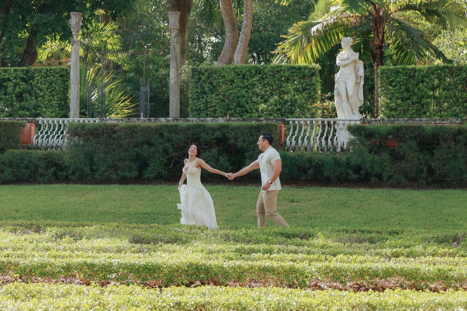 A couple holds hands while walking through a manicured garden with classical statues and palm trees.
