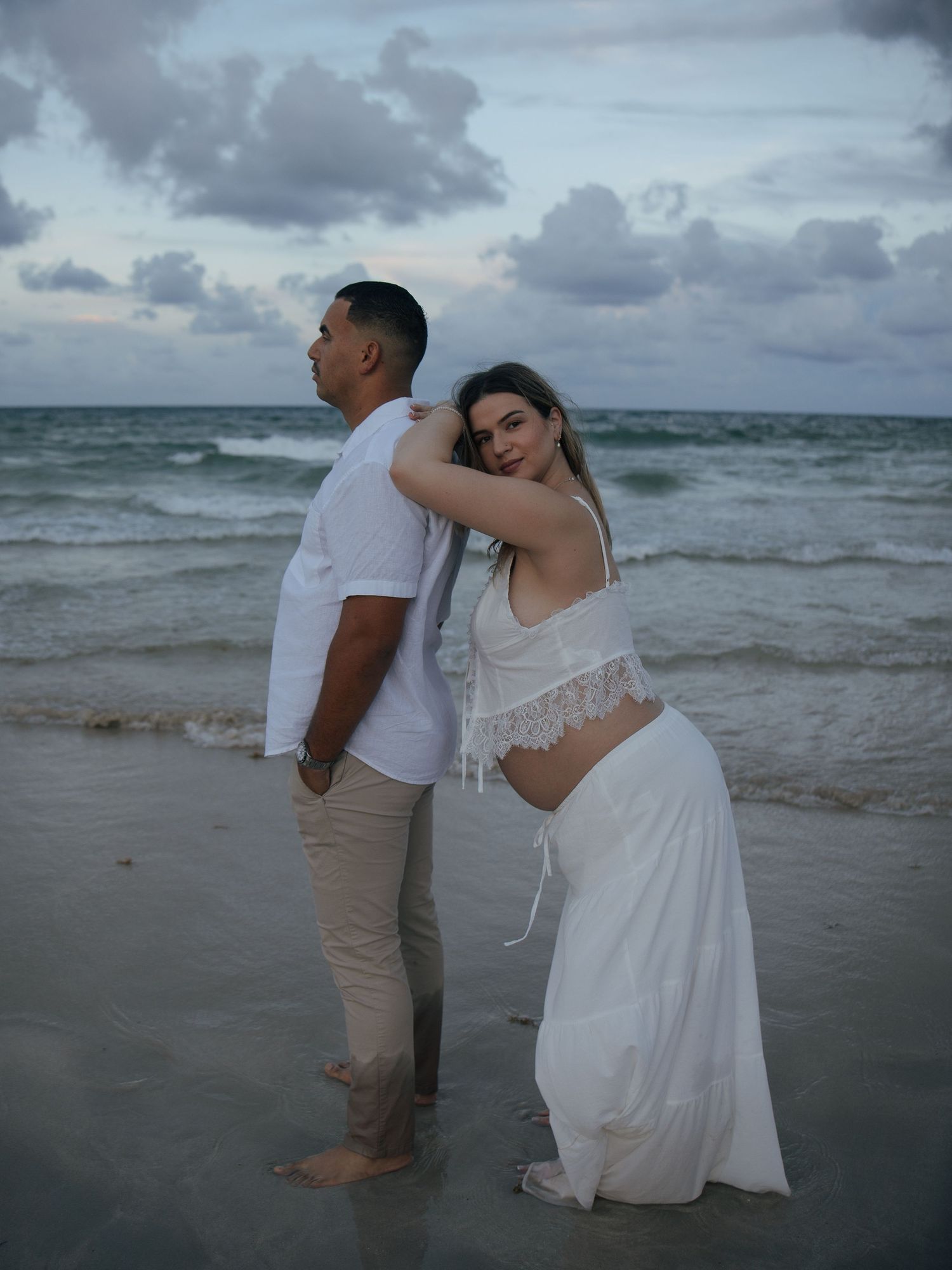 Pregnant woman in white dress leaning on partner's back during beach maternity photoshoot at sunset.