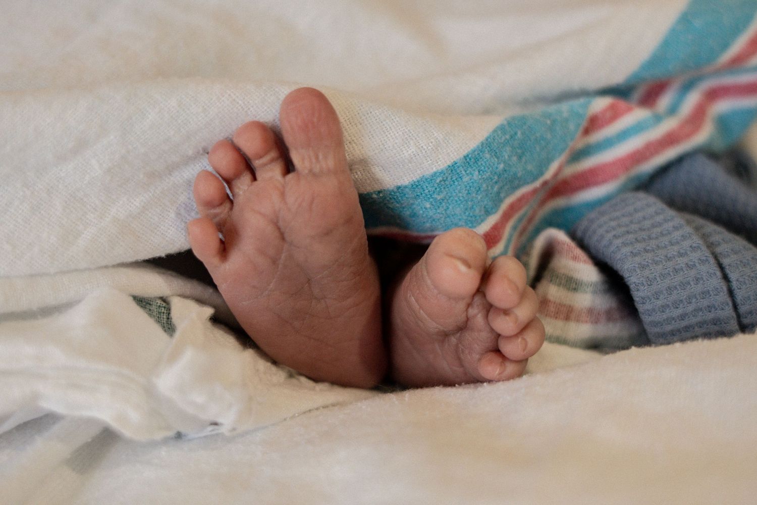 Tiny newborn baby feet peeking out from colorful striped blanket on white hospital bed.