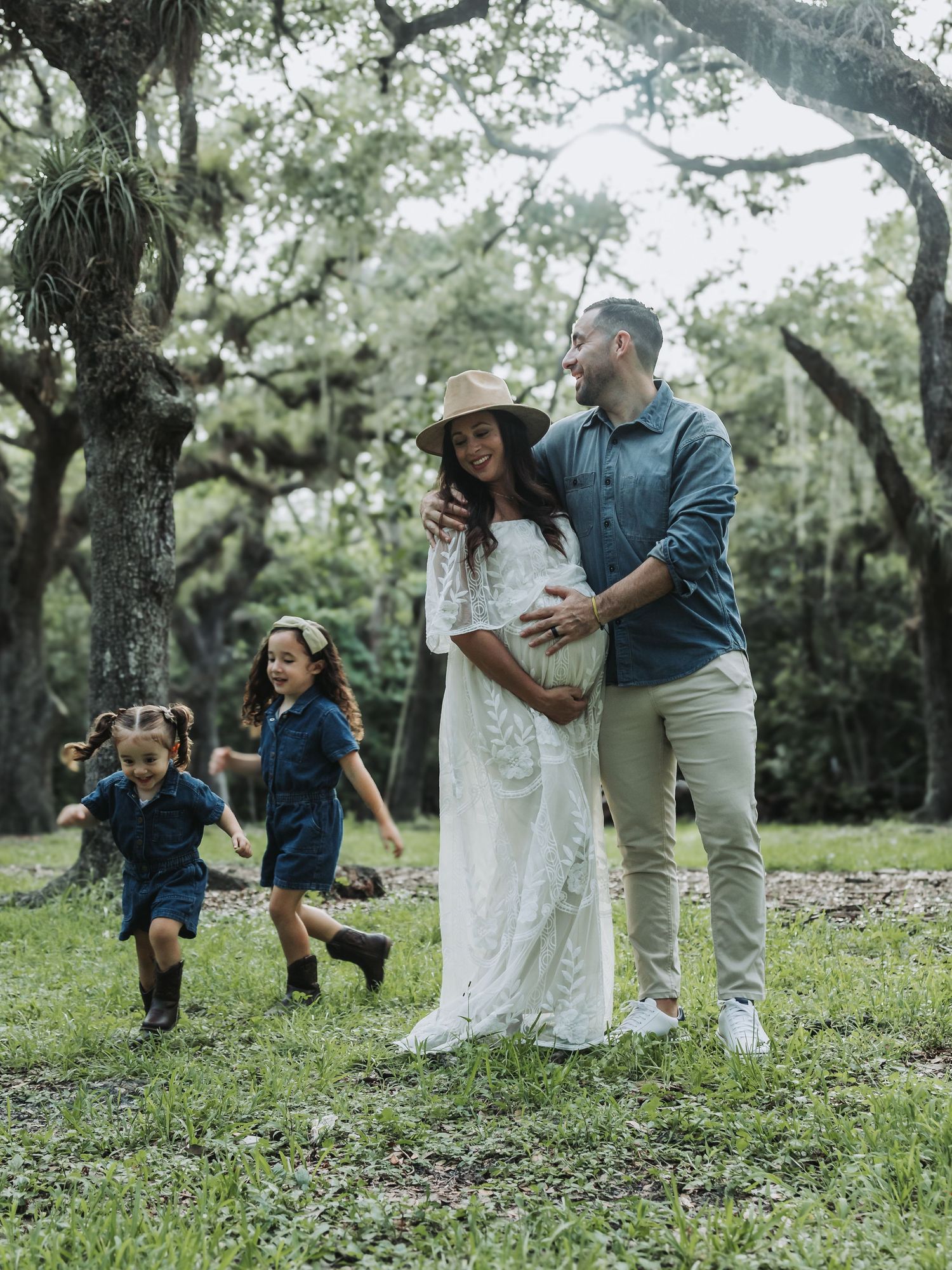 A couple cradles a baby bump while their two daughters play around them in a green park.