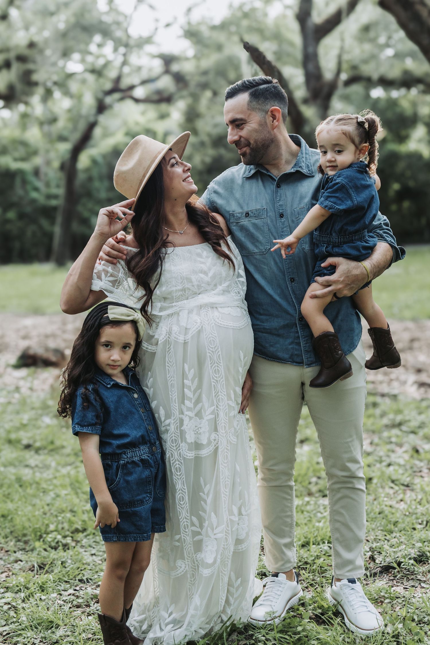 A pregnant mother and father smile together with their two young daughters in a lush green park.