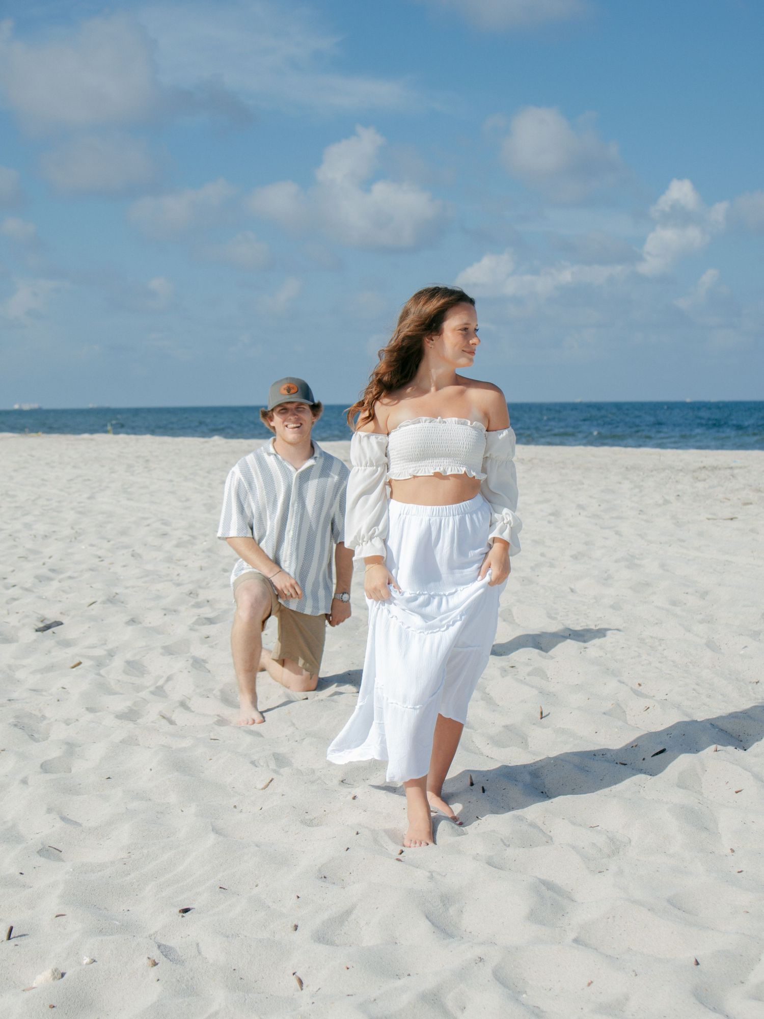 A young couple walks on a white sandy beach under a blue sky, dressed in coordinating white outfits.