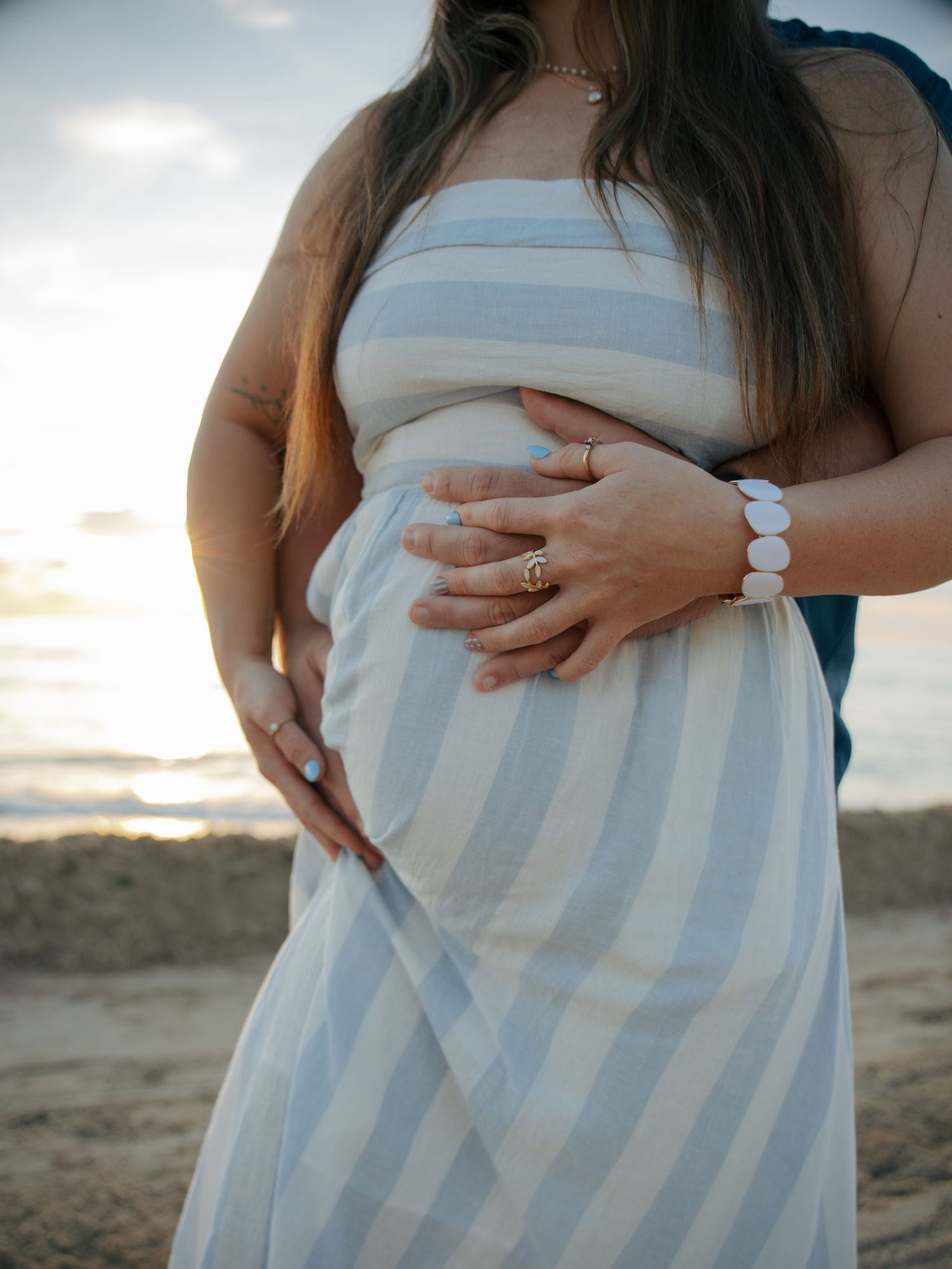 Close-up of pregnant woman in striped dress with partner's hands cradling baby bump at beach sunset.