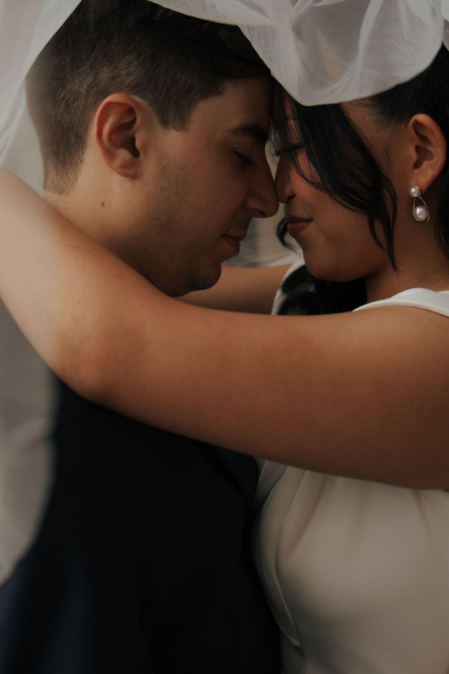 Intimate close-up of bride and groom touching foreheads under wedding veil with eyes closed.