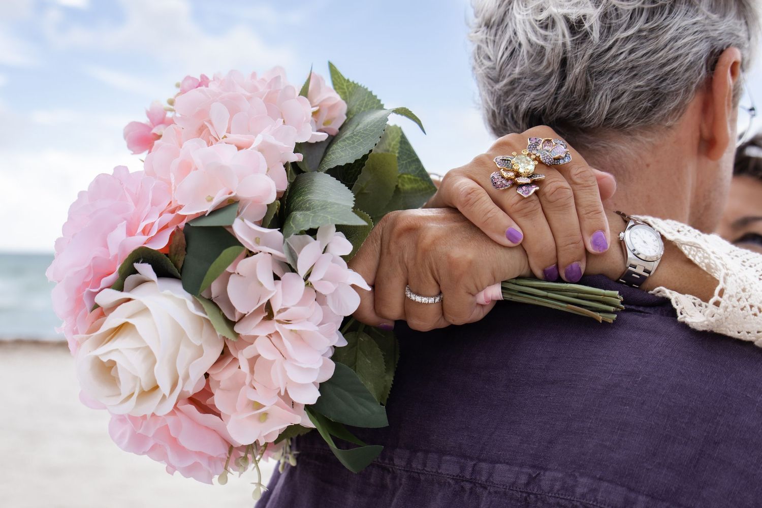 Woman holding pink and white flower bouquet around mans neck on a beach, wearing rings and watch.