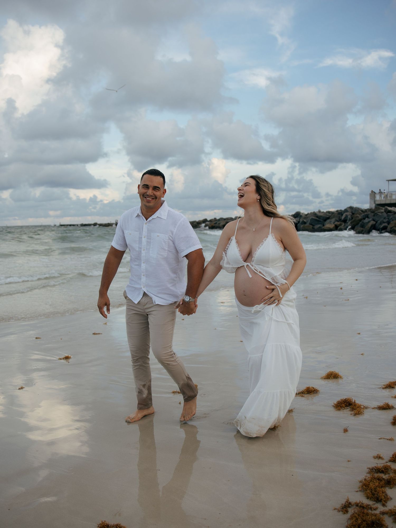 Happy expecting couple walking hand-in-hand on beach, woman in white maternity dress holding baby bump.