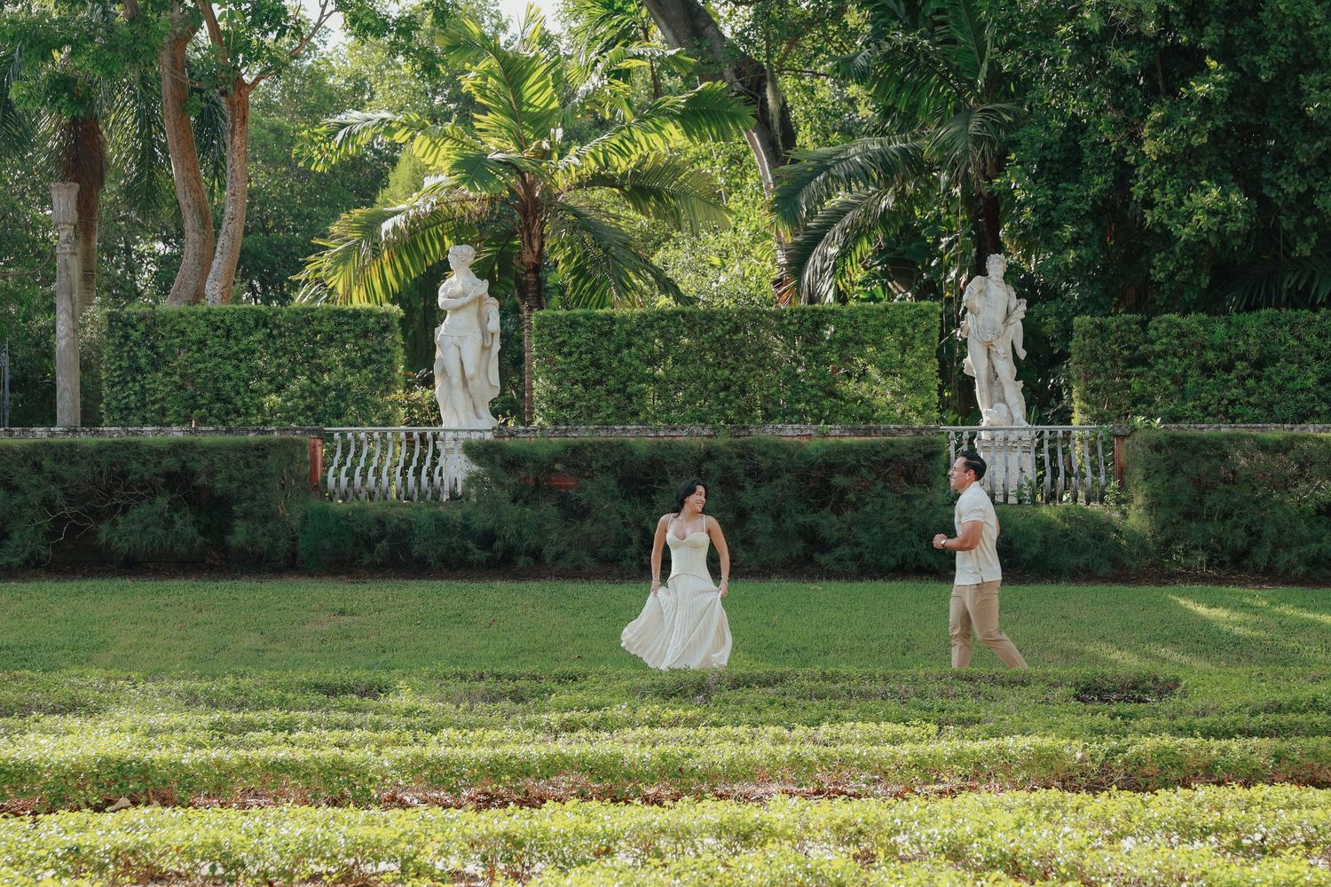 A couple strolls through a lush formal garden with sculpted hedges and classical stone statues.