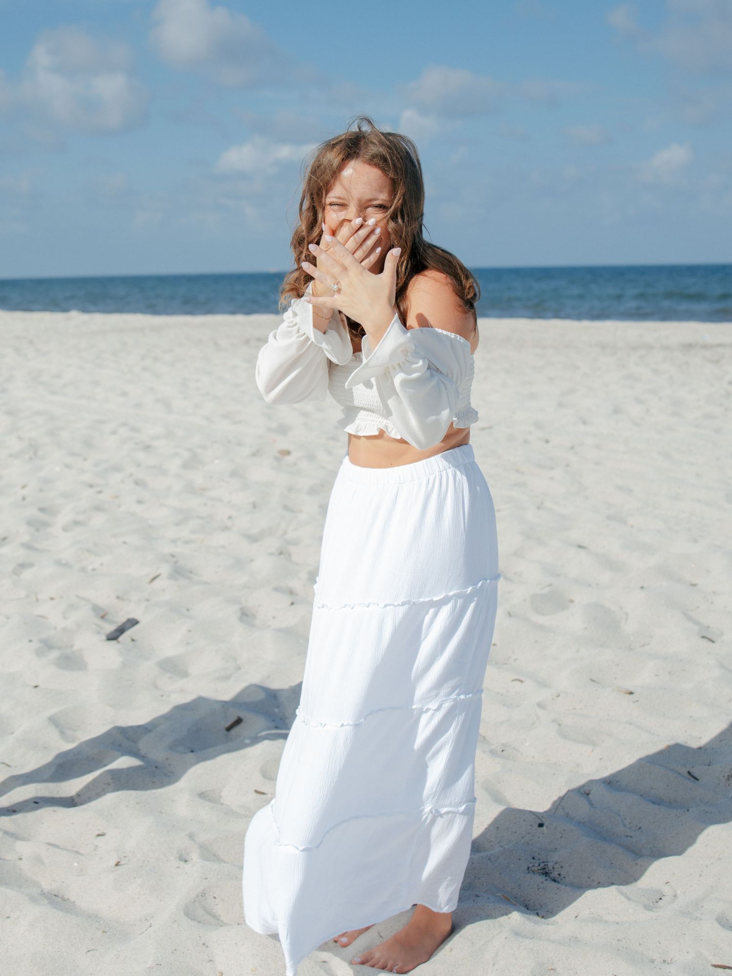 Surprised woman in white off-shoulder crop top and maxi skirt covering mouth with hands on sunny beach.