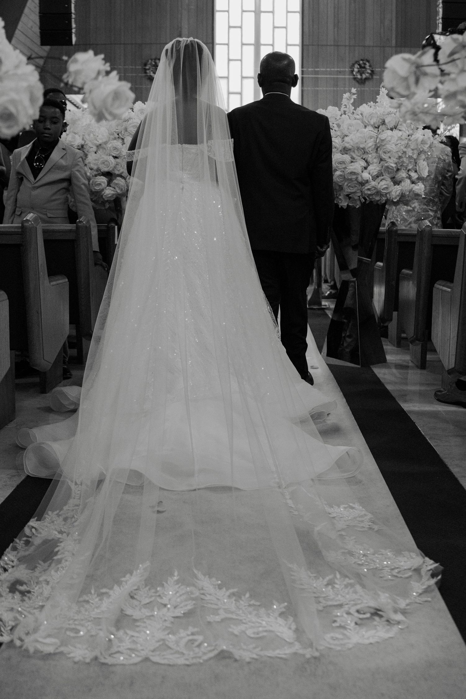 Black and white photo of bride walking down church aisle with long lace cathedral veil.