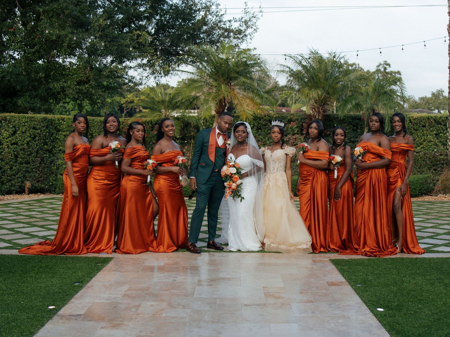 Wedding party poses outdoors with bridesmaids in burnt orange gowns and groomsmen in dark green suits.