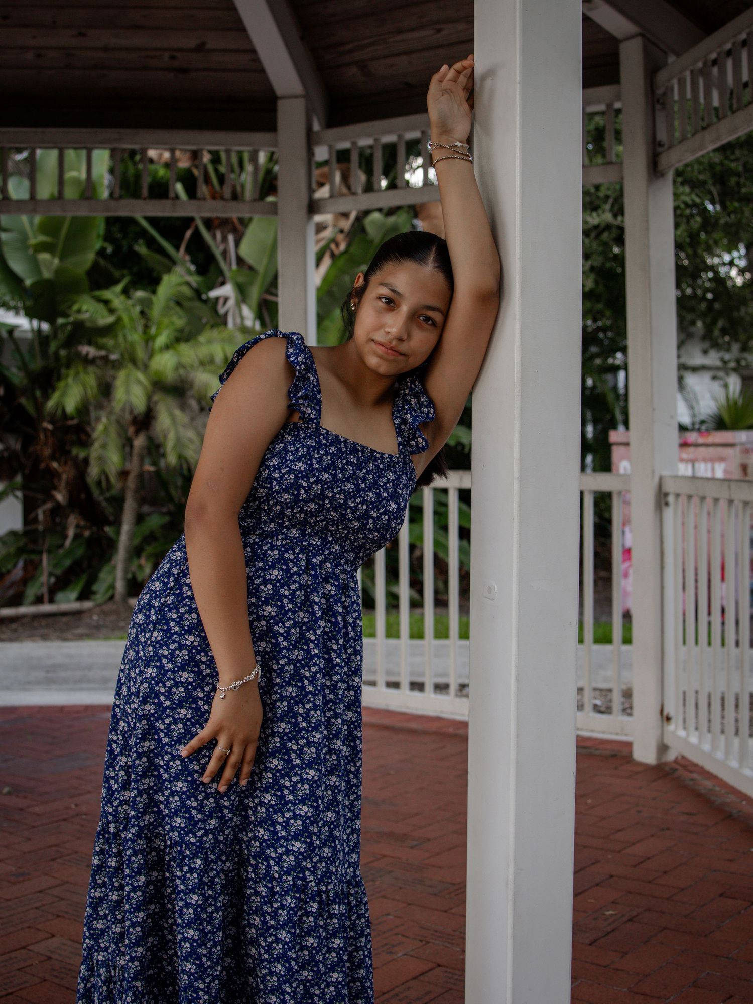 Teen girl in blue floral dress leaning against gazebo column with arm raised overhead.