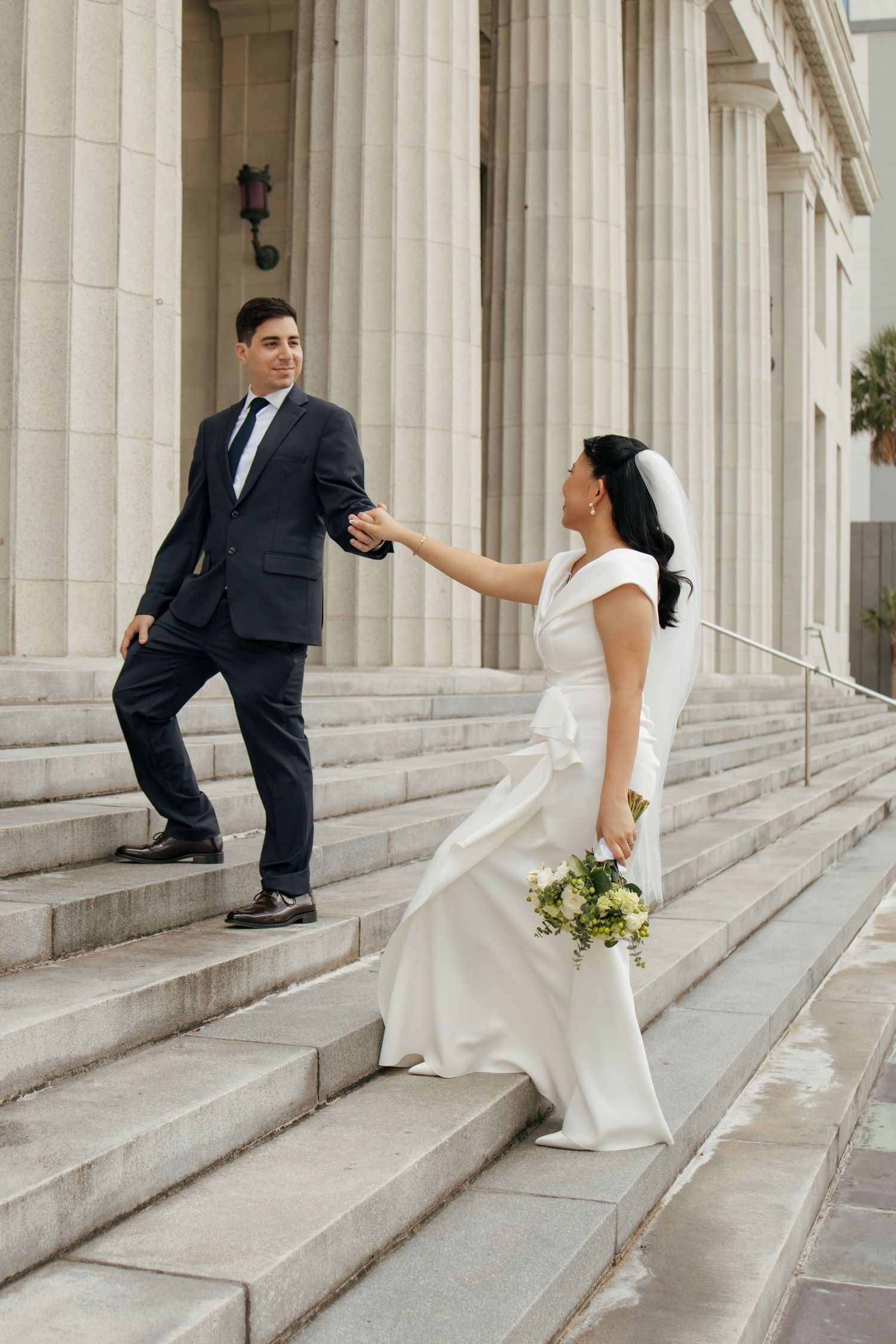 Bride in white gown holding bouquet takes groom's hand on grand stone courthouse steps.