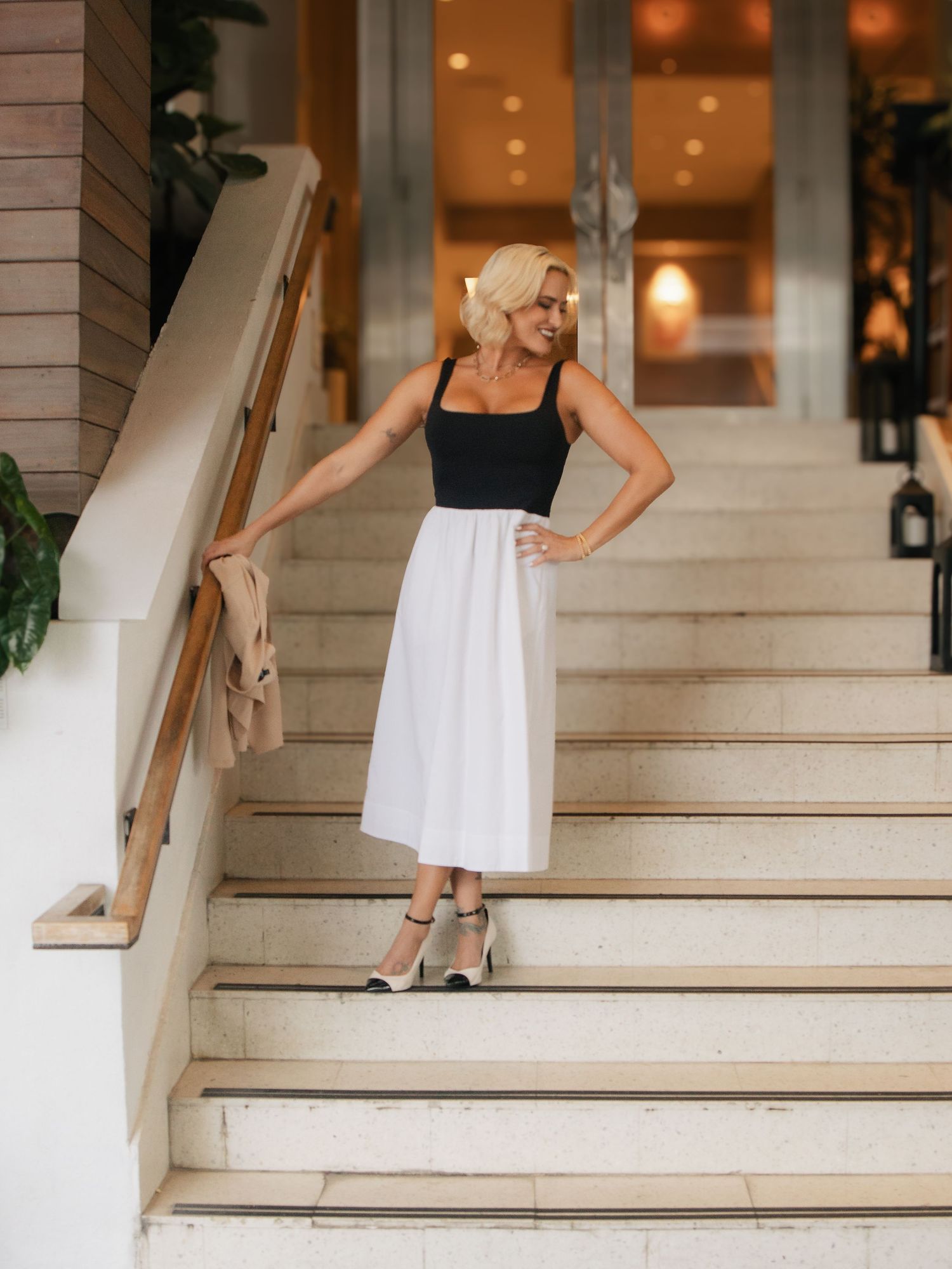Blonde woman in black tank top and white midi skirt posing on marble staircase in elegant venue.