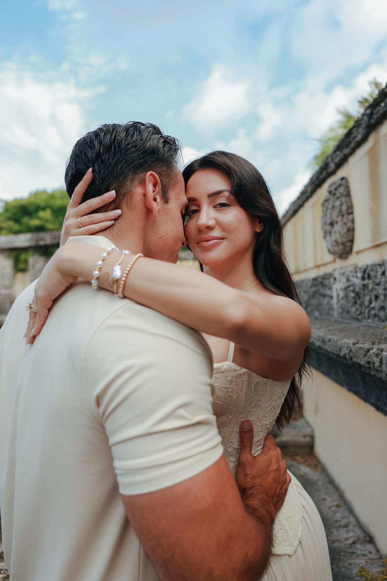 A woman with dark hair smiles while embracing a man outdoors under a cloudy blue sky.
