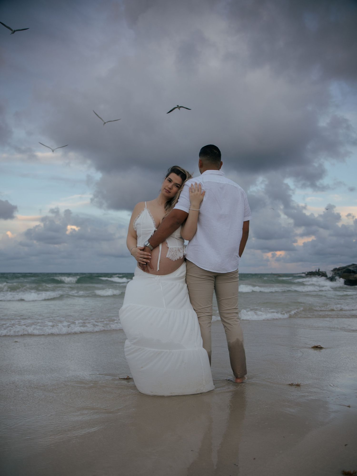 Pregnant couple in white outfits embracing on a beach at dusk with dramatic cloudy sky and ocean waves.