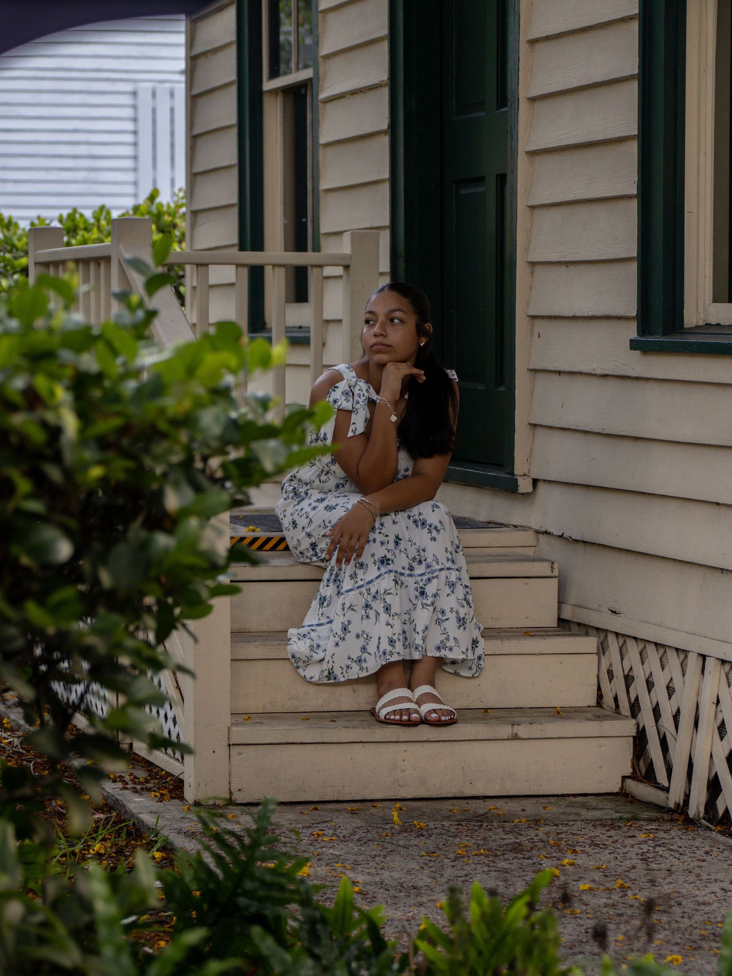 Thoughtful young woman in white floral skirt sitting on porch steps beside lush greenery.