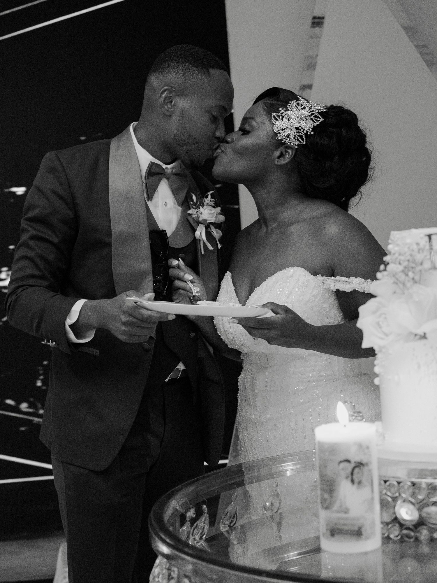 Black and white photo of a couple sharing a kiss while cutting their wedding cake at their reception.