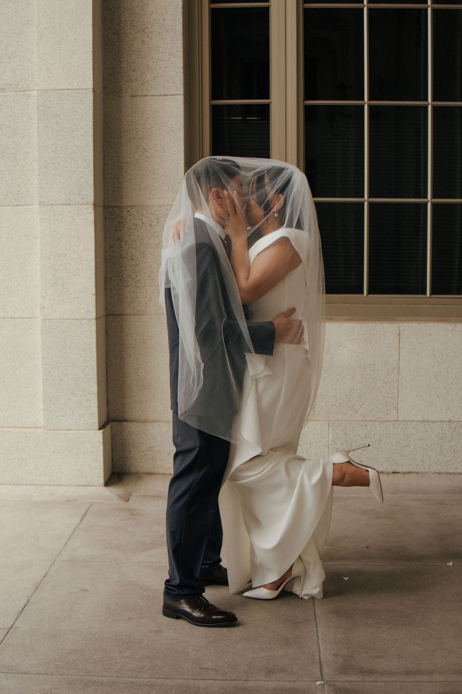 Bride and groom sharing a kiss under flowing veil against stone building exterior.