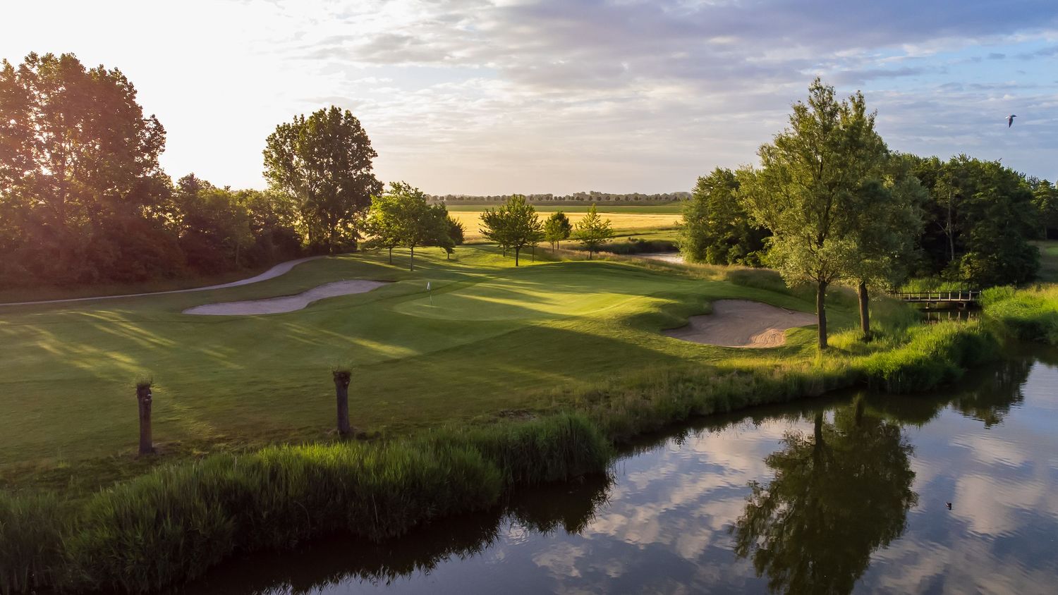 Sunlit golf course fairway with sand bunkers beside a calm reflective water hazard.