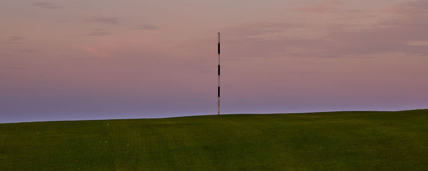 Lone golf flag pin standing on a green hill against a soft pink dusk sky over the horizon.