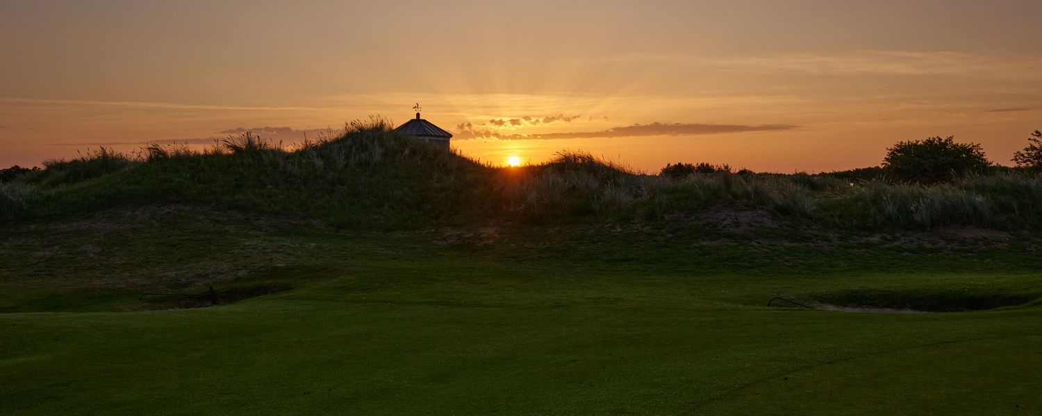 Dramatic orange sunset silhouetting a links golf course hole with sandy dunes and green fairway.