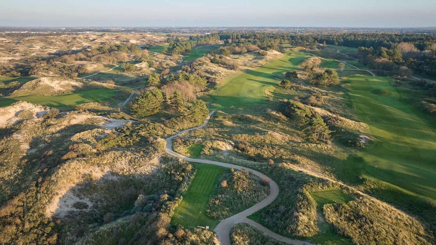 Aerial view of a links golf course with rolling dunes, winding cart paths, and lush green fairways at golden hour.