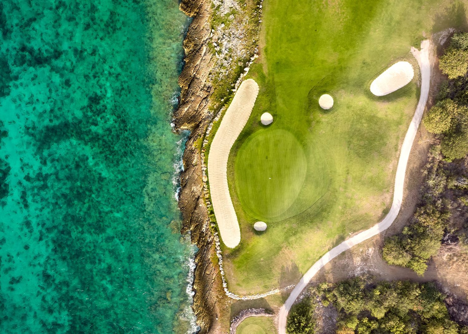Aerial view of coastal golf course green with sand bunkers beside turquoise ocean waters.