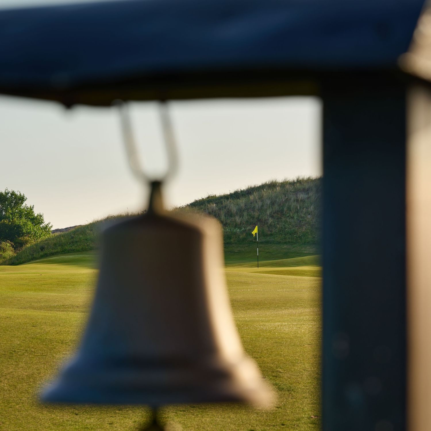 Bronze bell in foreground frames a golf green with yellow flag amid rolling dunes at sunset.