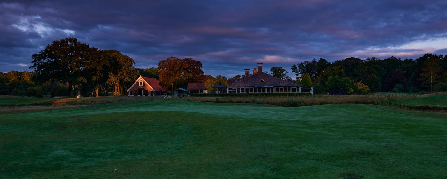 Golf course green at dusk with illuminated clubhouse and dramatic purple sky backdrop.