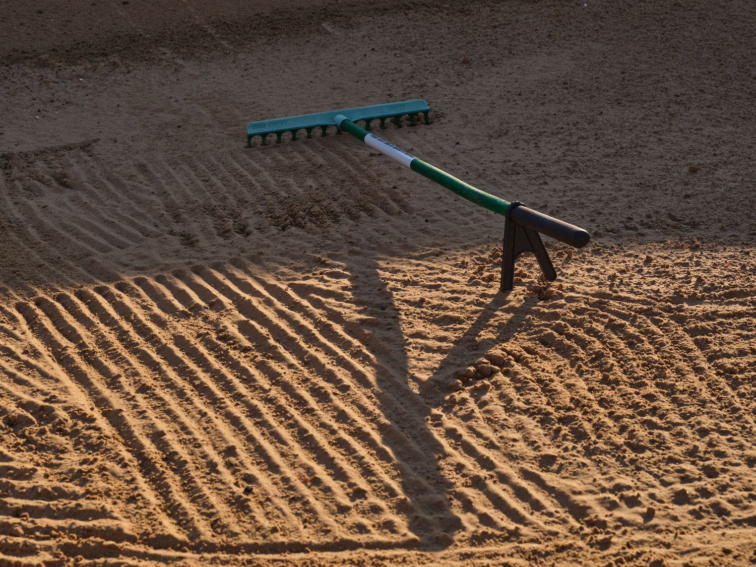 Sand bunker raked in neat parallel lines with a green rake casting a long shadow in bright sunlight.