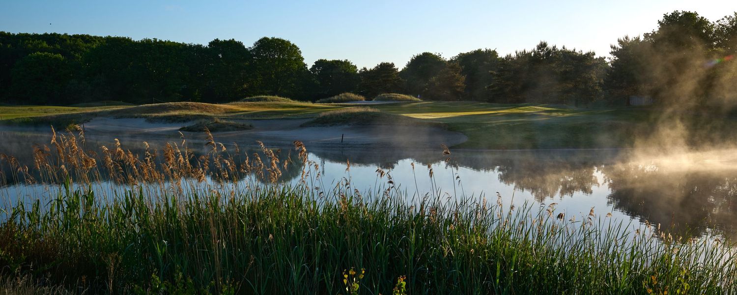 Misty morning golf course with fog rising over a serene pond surrounded by tall grasses and trees.