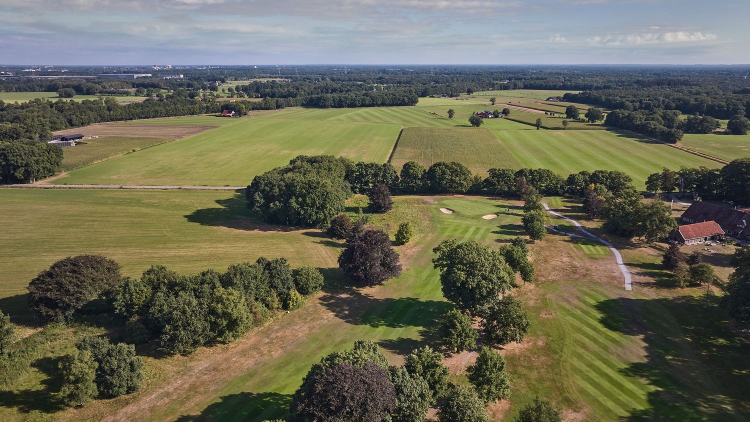 Aerial view of a lush green golf course surrounded by trees and farmland under a clear blue sky.