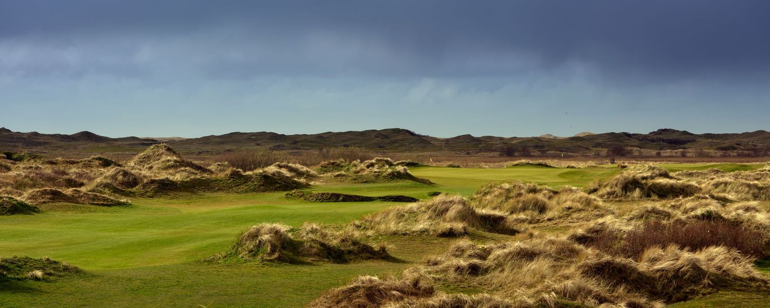 A links golf course fairway winds through rugged sand dunes under a dramatic stormy sky.