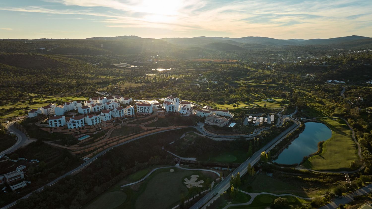 Aerial sunset view of a luxury golf resort with a lake, white buildings, and rolling green course.